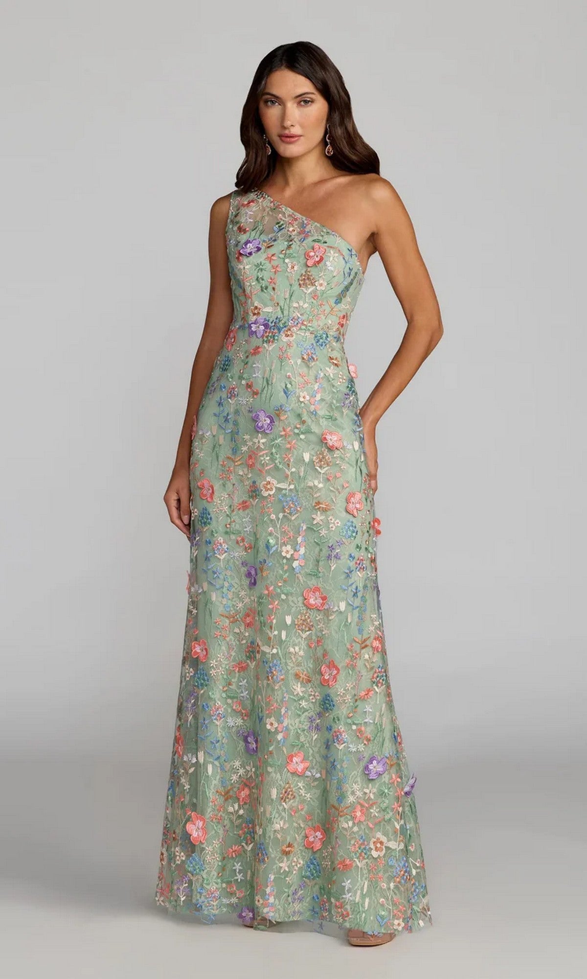 A woman wears the Mac Duggal 68606 long floral-print formal dress, featuring a one-shoulder design, standing against a plain gray background.